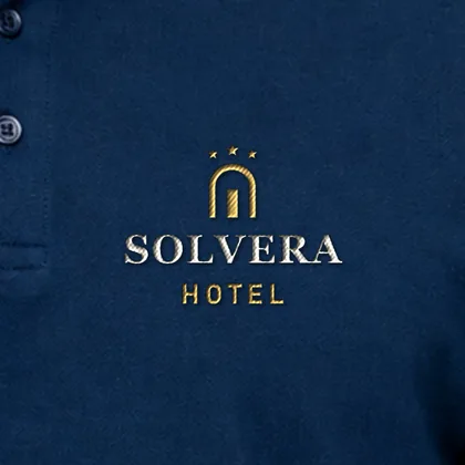 Zbliżenie haftu przykładowego logo hotelu SOLVERA na granatowej koszulce polo — złota i kremowa nić, widoczna dzianina pique (logo wymyślone, zdjęcie poglądowe)